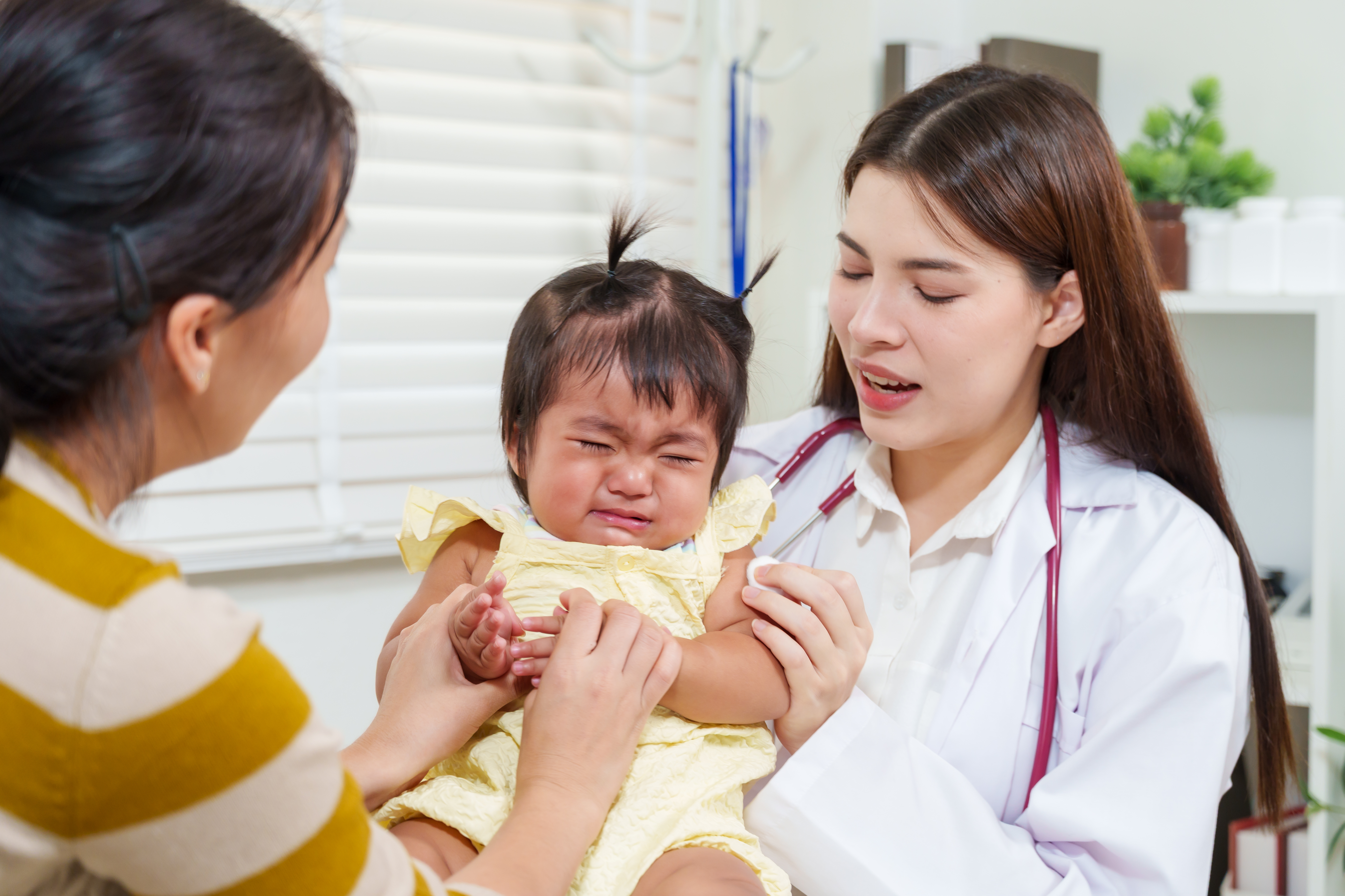 Asian mother holding crying toddler while pediatrician examining injection area after vaccination inside clinic showing empathy care and family support promoting trust in pediatric healthcare