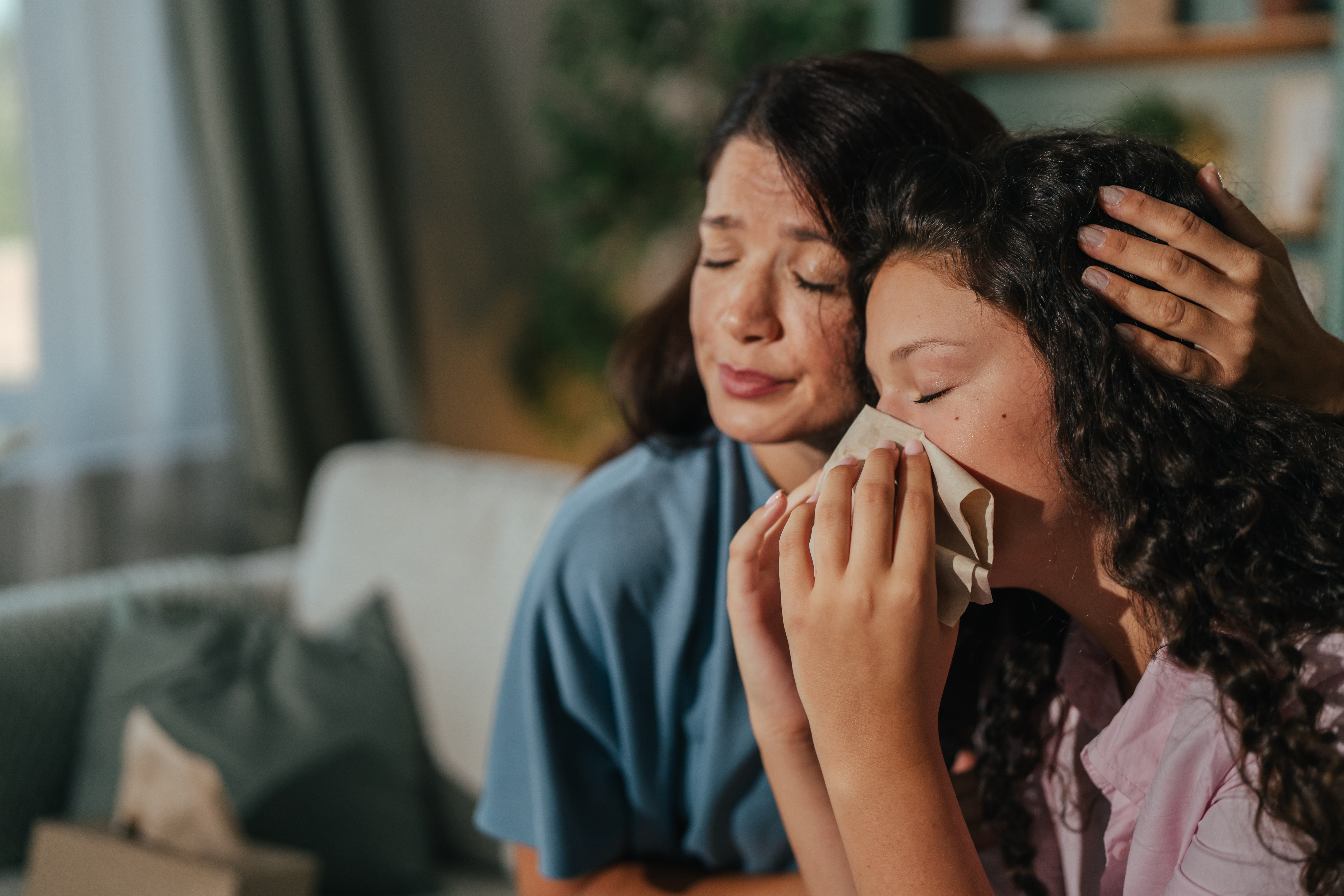 Mother providing comfort to her sad teenage daughter, gently placing a hand on her head while the girl wipes tears with a tissue, reflecting a moment of emotional support and connection